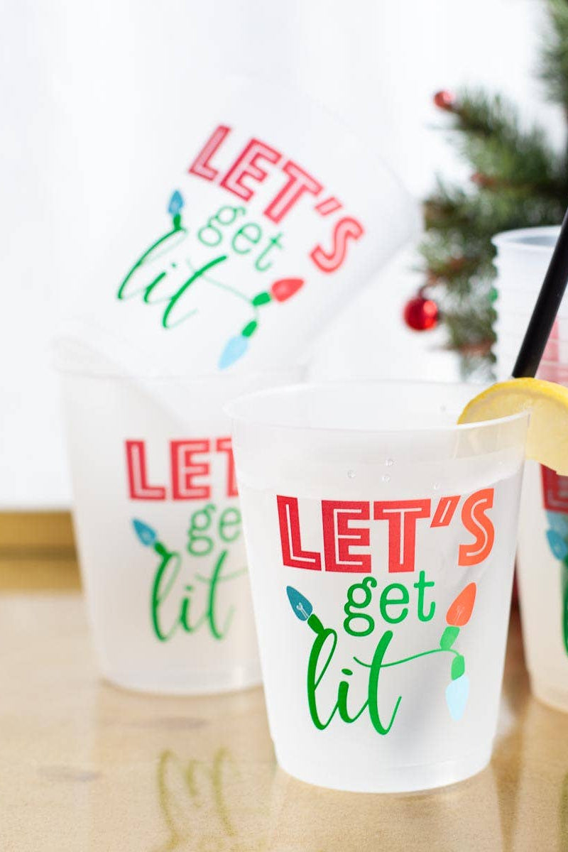 Clear plastic cups with 'LET'S get lit' text and Christmas lights design, on a wooden surface with a blurred Christmas tree in the background.